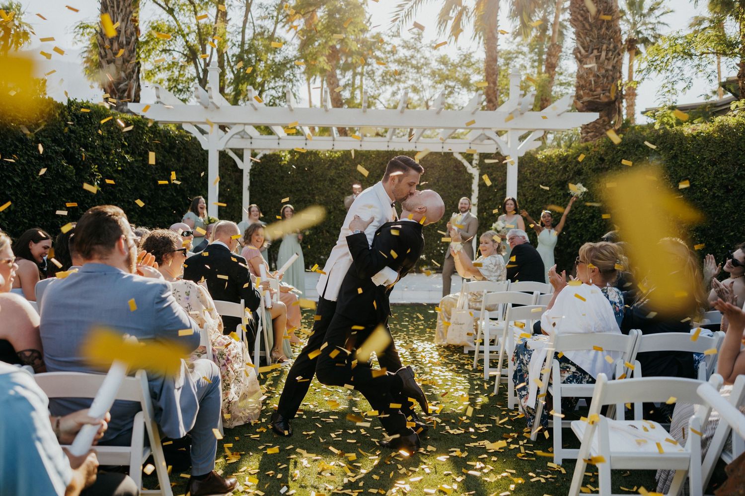 A jubilant outdoor wedding ceremony under a white pergola with sunlight streaming through the trees.