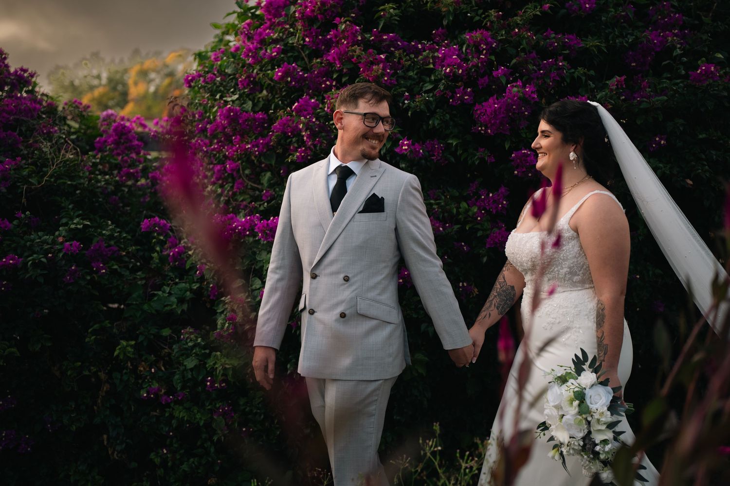 Bride and groom hold hands among vibrant purple bougainvillea in romantic outdoor wedding portrait.