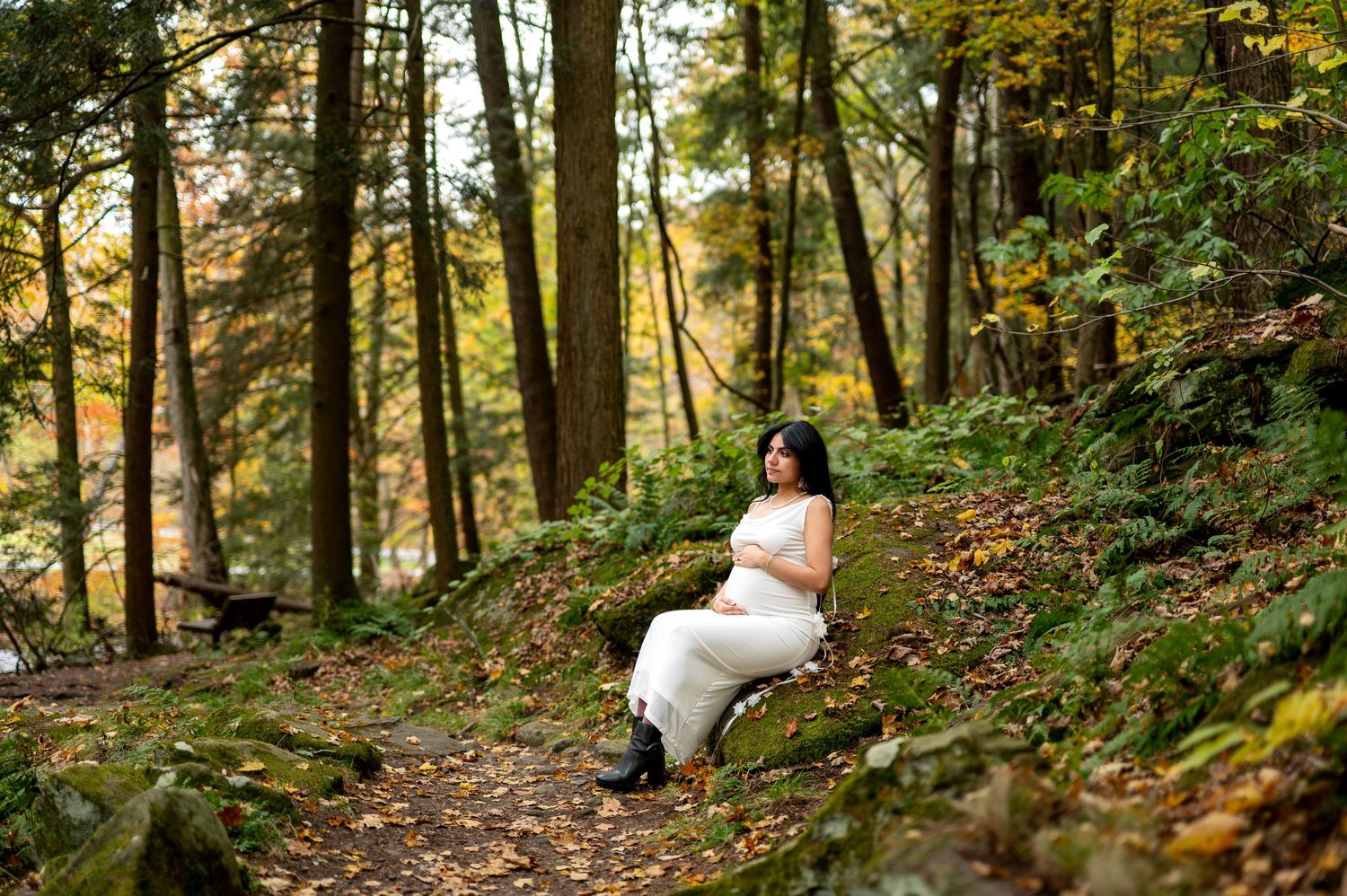 Pregnant woman in white dress sitting on a mossy rock in a serene autumn forest setting.
