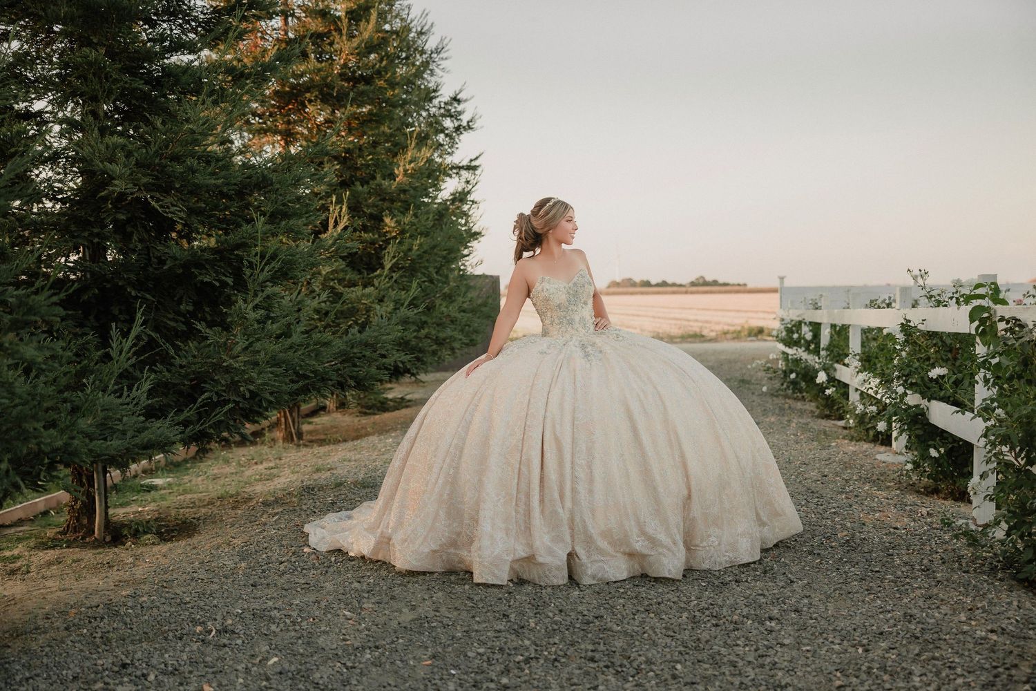 A quinceanera in a flowing champagne-colored ballgown stands on a gravel path at sunset for Bella Vista Farms quince celebration