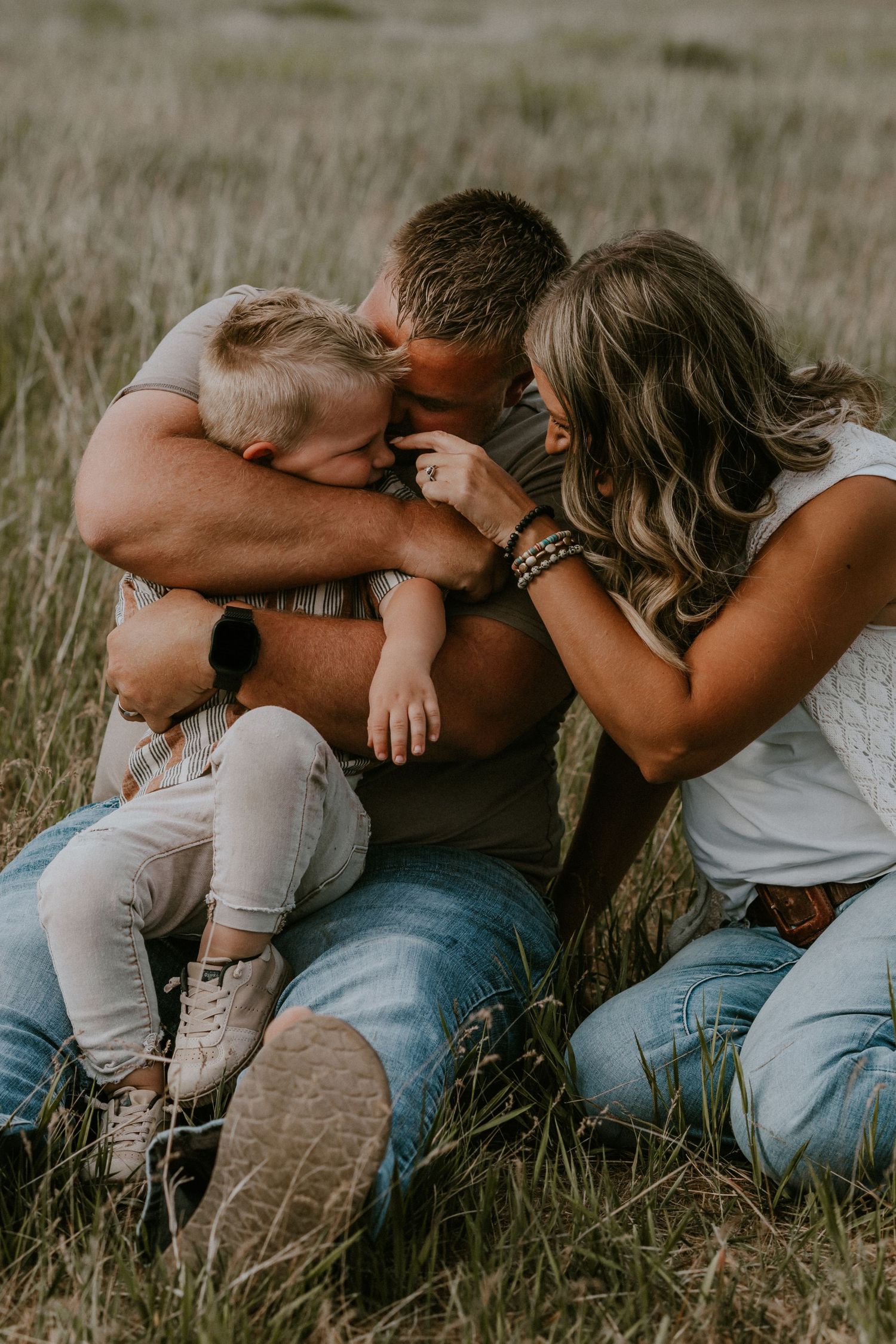 A couple in casual clothing lies in tall grass during an intimate outdoor photoshoot.