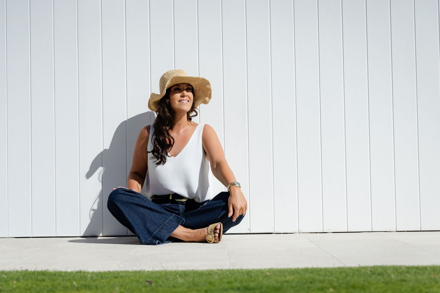 Woman in sun hat and white tank top sits cross-legged against white wall on grass in sunlight.