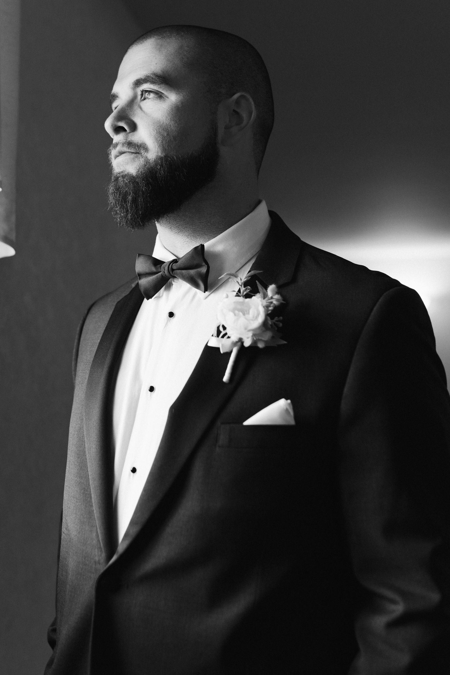 Black and white portrait of a groom in a tuxedo with boutonniere looking to the side in dramatic lighting.