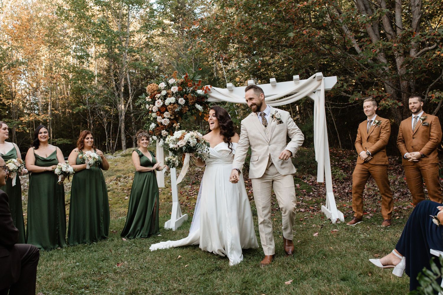 A wedding party poses in a wooded outdoor setting with bridesmaids in dark green dresses and groomsmen in brown suits.