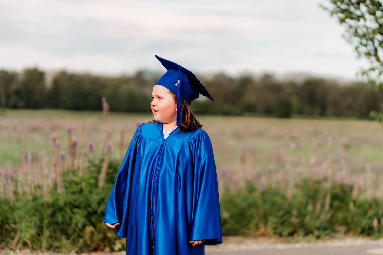 A person in bright blue graduation regalia stands in a wildflower field during a cap and gown photo session in Puyallup.