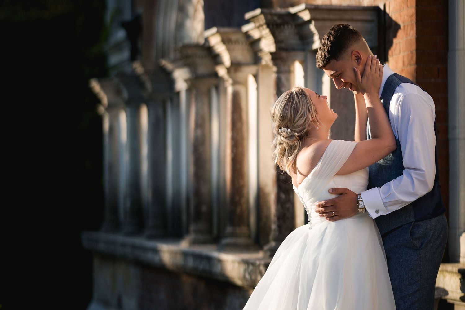 Romantic couple in wedding attire share intimate embrace in sunlit stone archway corridor.