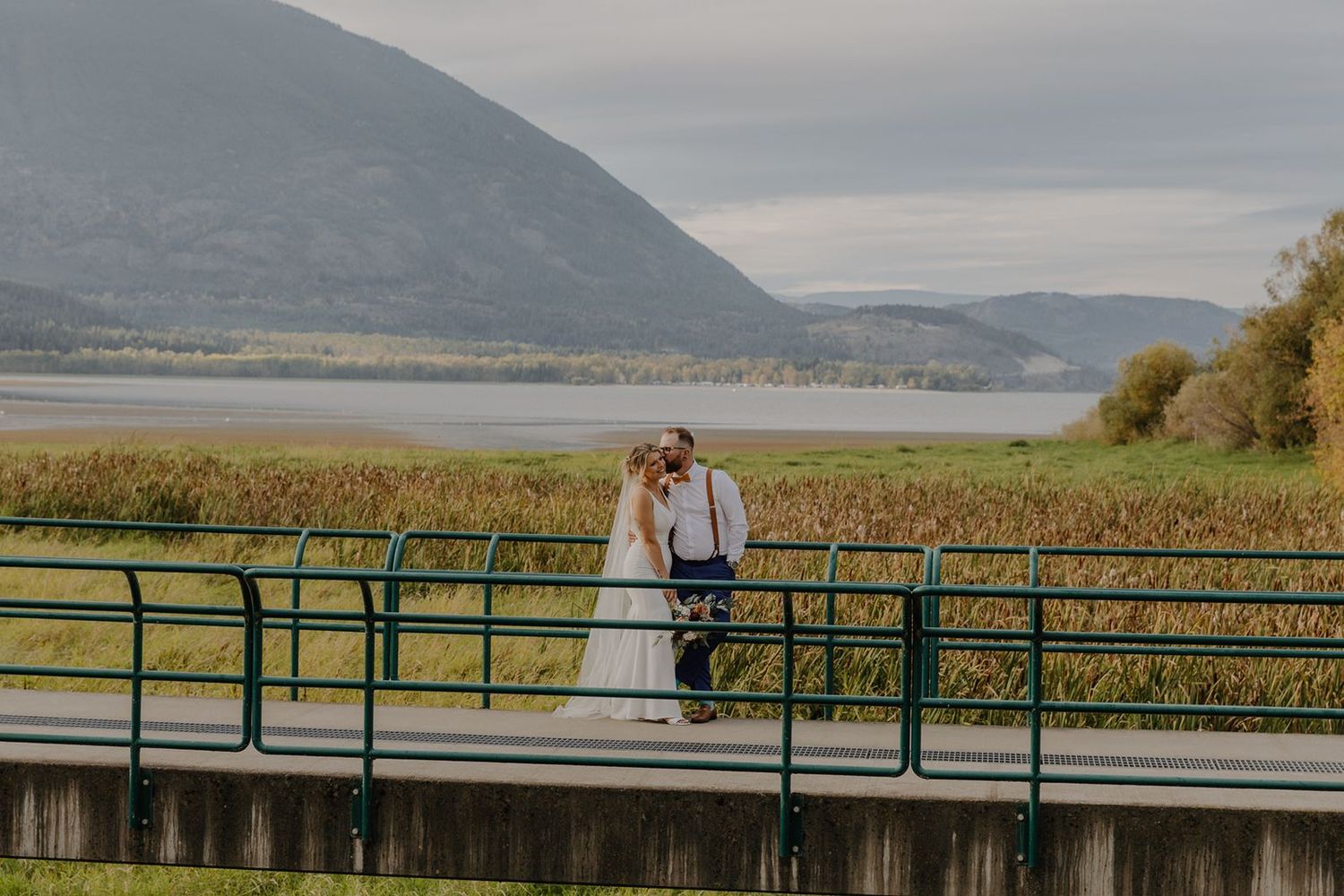 Bride and groom sharing a kiss on a bridge with scenic mountain lake views on their wedding day.