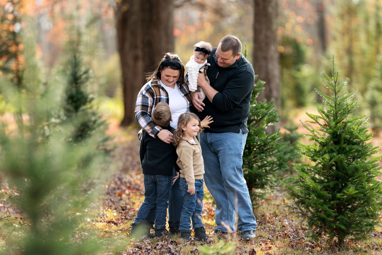 A family of five shares a playful moment together among small evergreen trees during an autumn outdoor photo session.