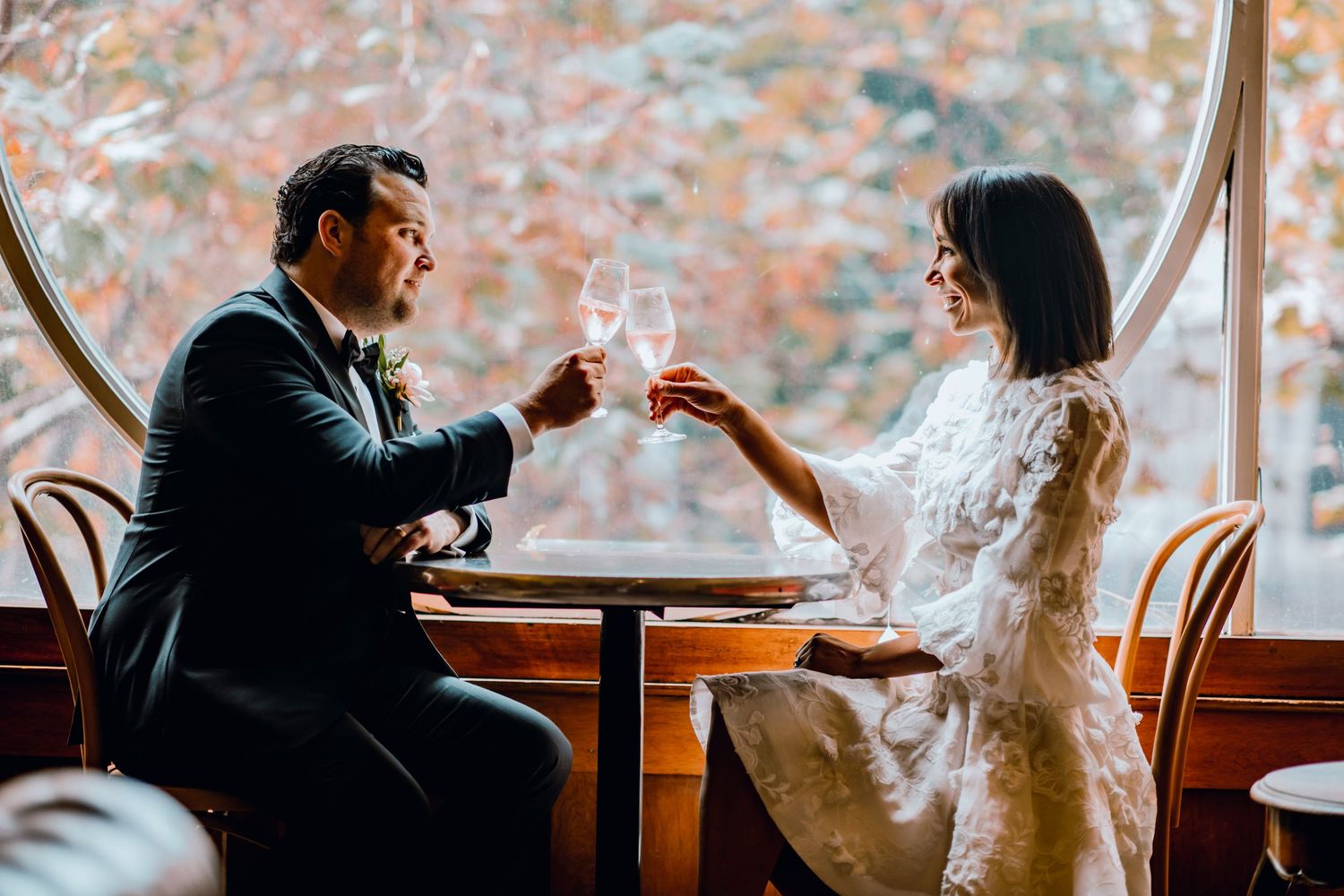Couple sharing an intimate moment during dinner at a restaurant with large windows overlooking an autumn landscape.