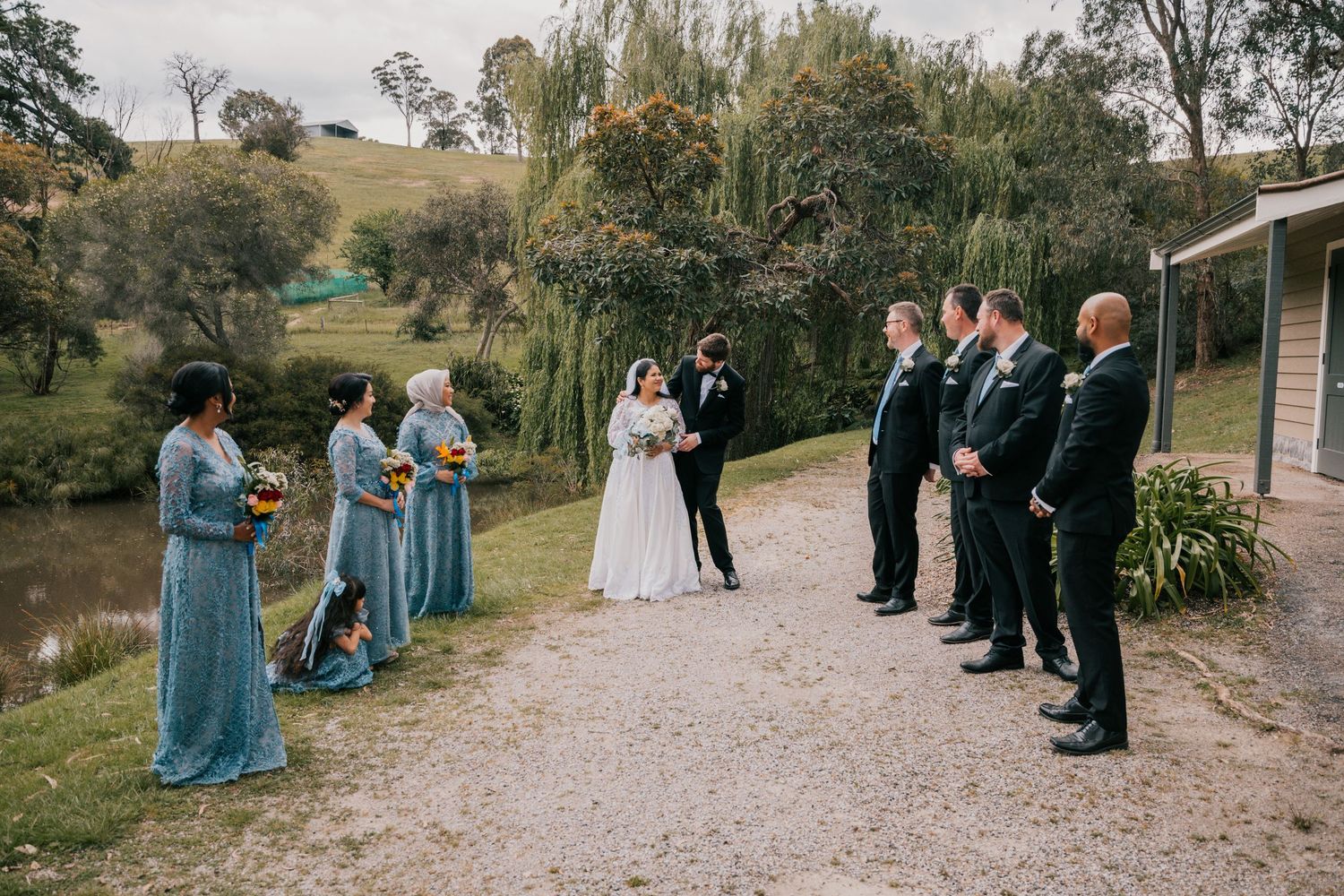 Wedding ceremony attendees gather outdoors on a gravel path lined with brick walls and autumn foliage.