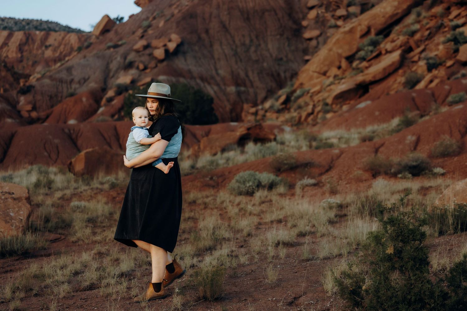 A woman in a black dress and hat holds her baby while standing among red rock desert formations.