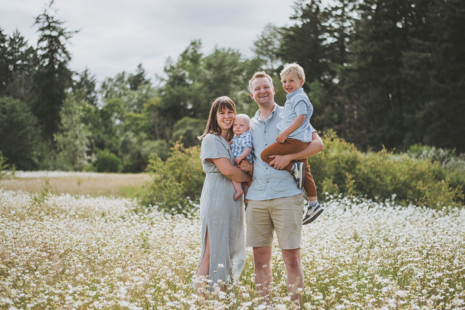 A family of four wearing coordinated blue and grey outfits embrace joyfully in a blooming wildflower daisy field on a sunny day.