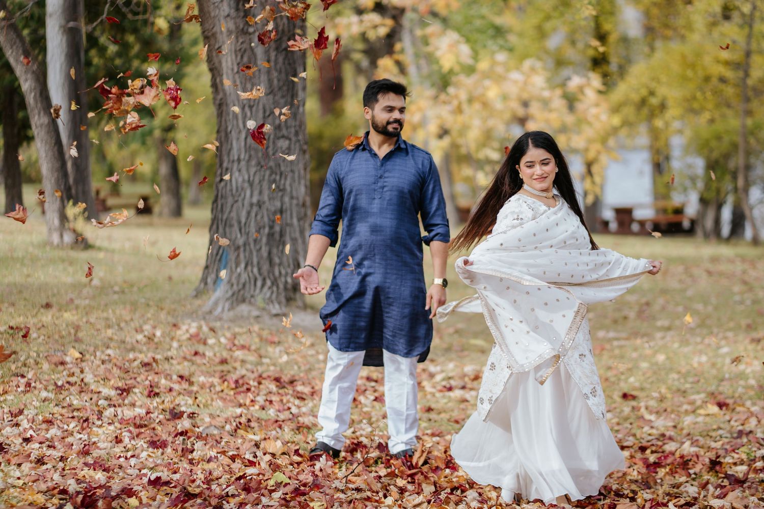 A couple in navy blue and white attire playfully throw autumn leaves in a park setting during a photo session.
