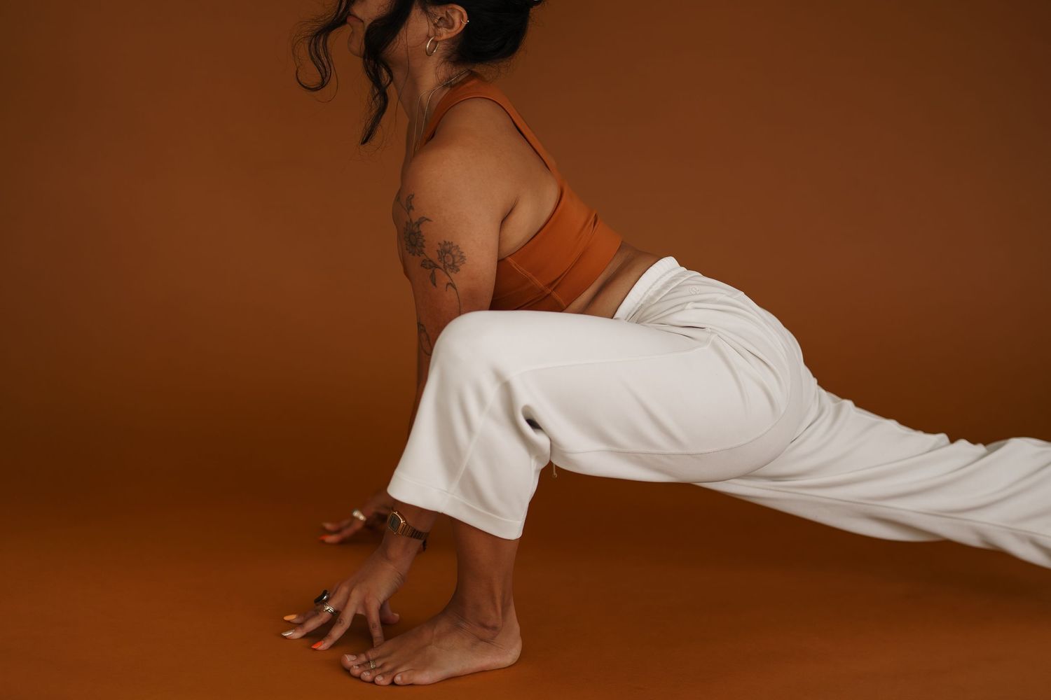 Athletic person wearing white pants and orange top stretches in low lunge yoga pose against brown backdrop.