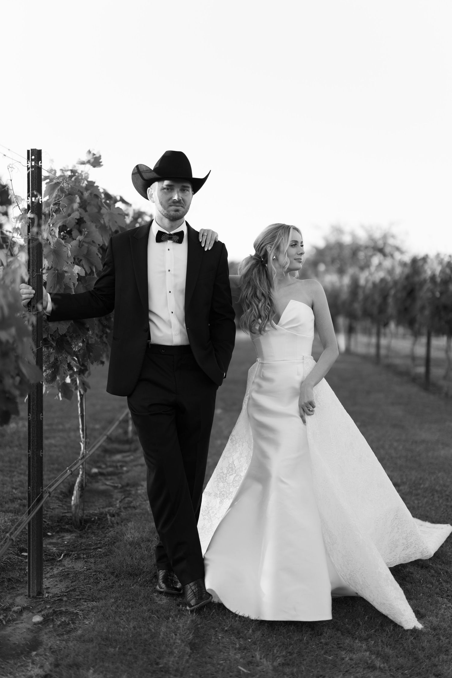 A newlywed couple poses playfully in a vineyard with the bride in white and groom in black tuxedo at sunset.