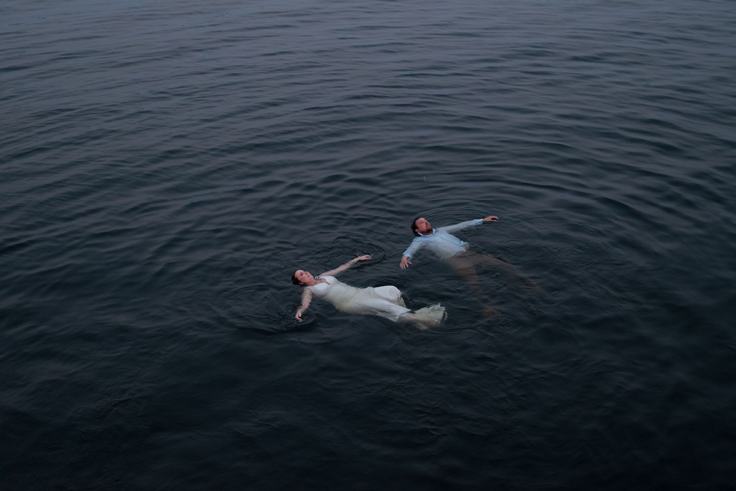 Two people floating and relaxing in dark ocean waters wearing white clothing and red swim caps.