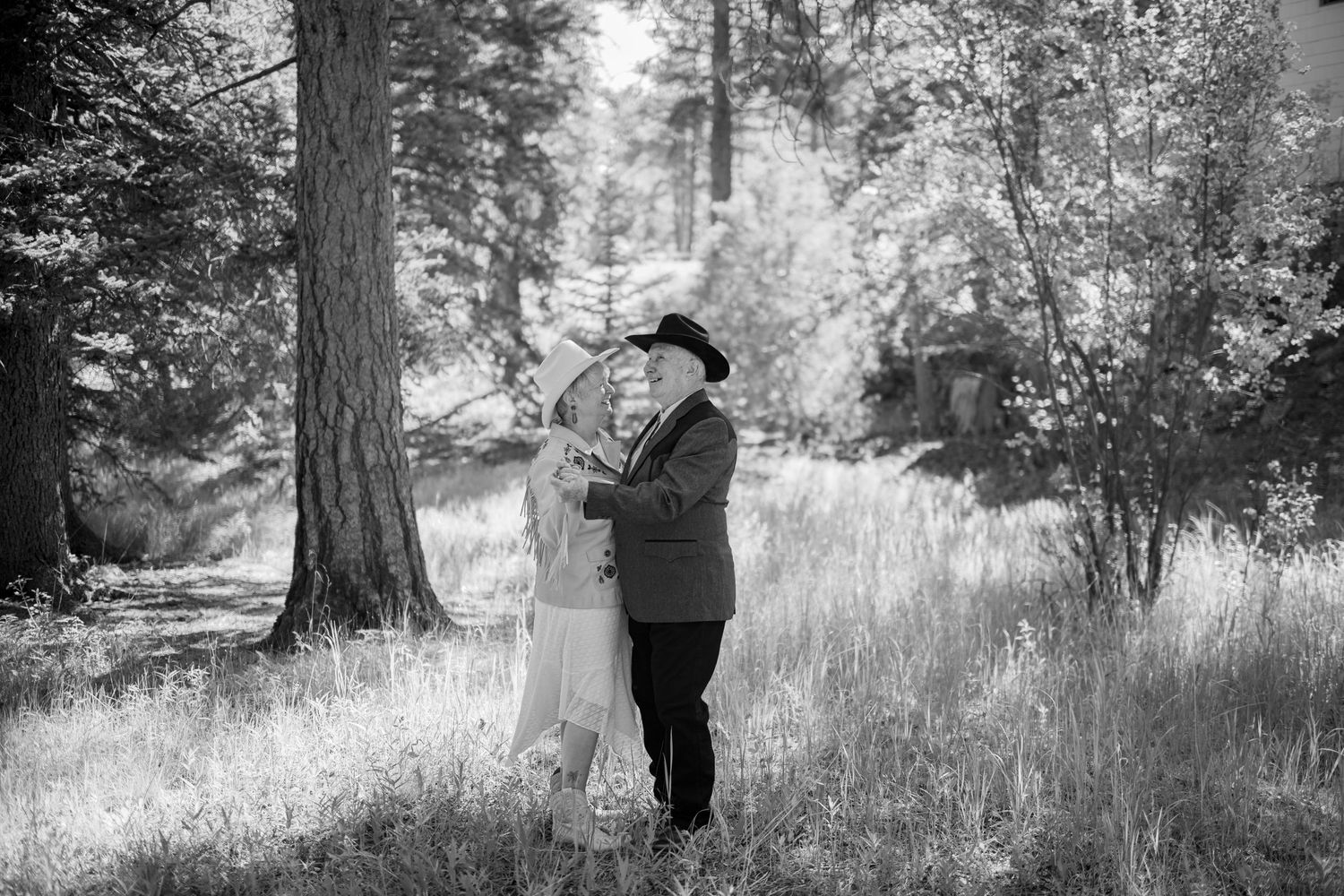 A romantic couple embraces in a sunlit forest clearing surrounded by tall trees in black and white photography.