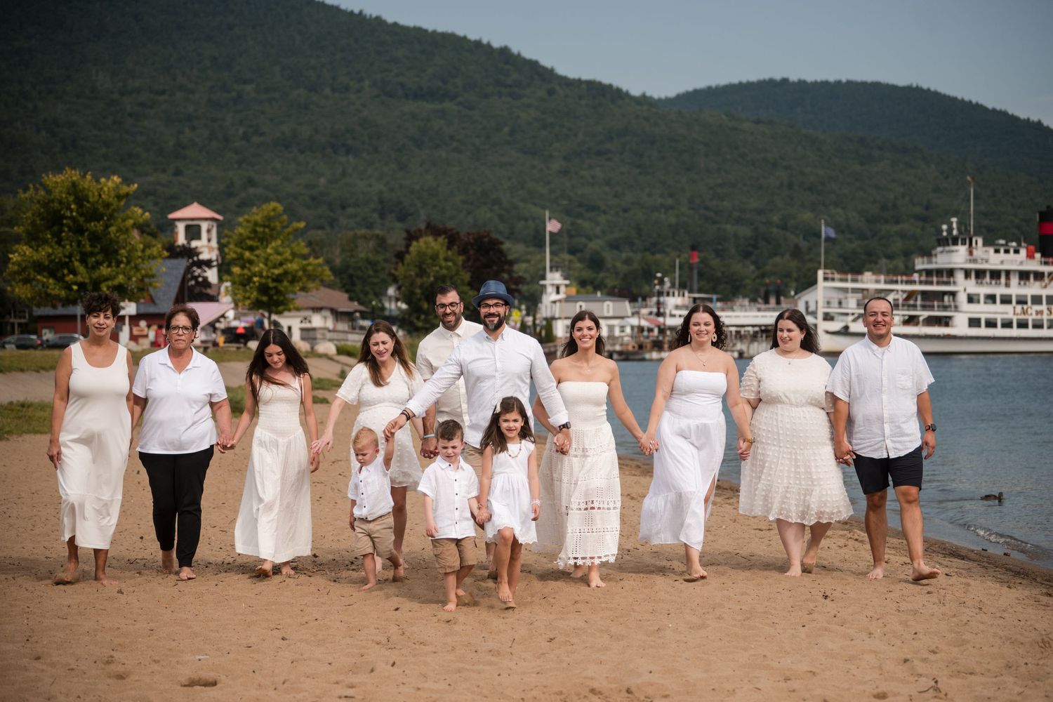 A group in coordinated white outfits poses together on a beach with mountains and a boat visible in the background.