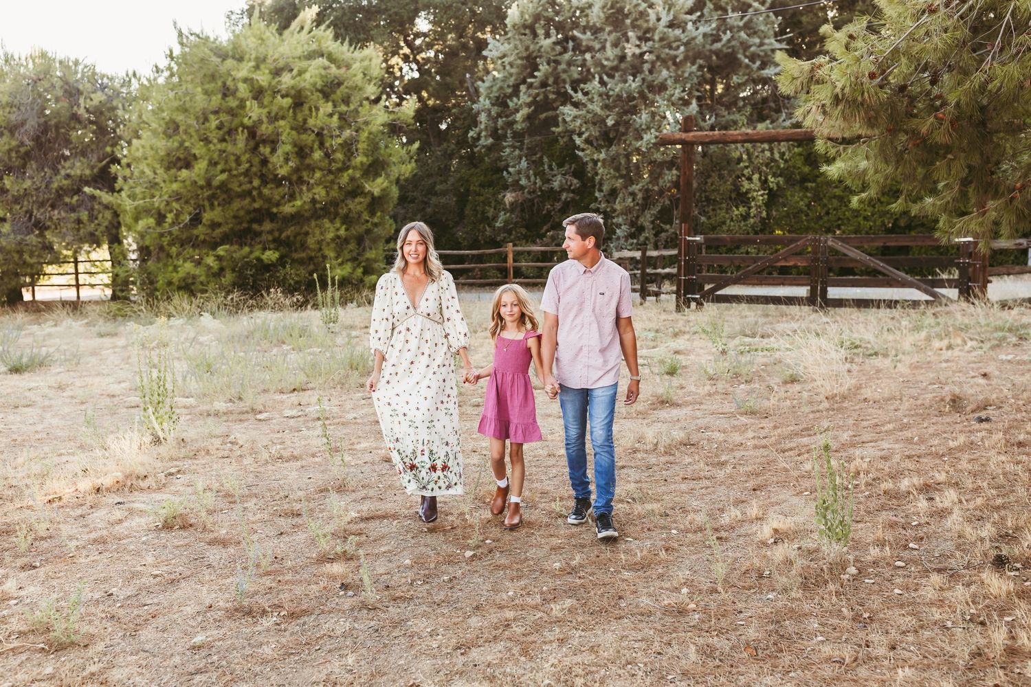 A family walks hand in hand along a dirt path with rustic wooden fencing and trees in the background.