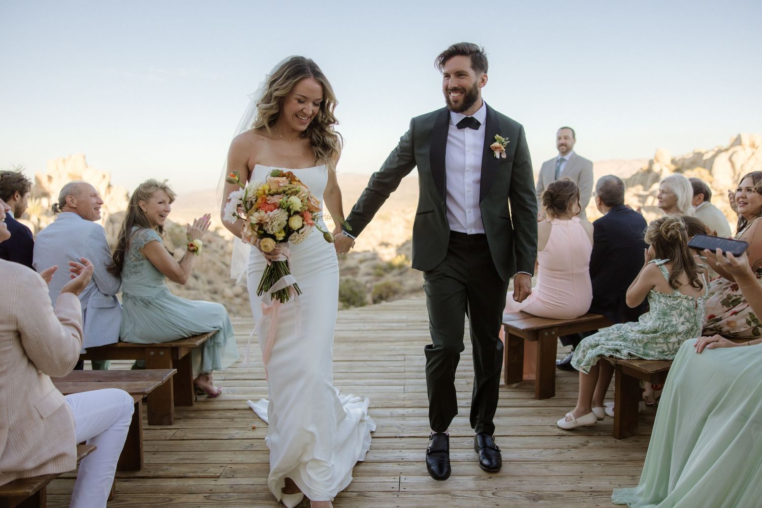 A joyful couple walks down the aisle at their outdoor wedding ceremony while guests celebrate from wooden benches.