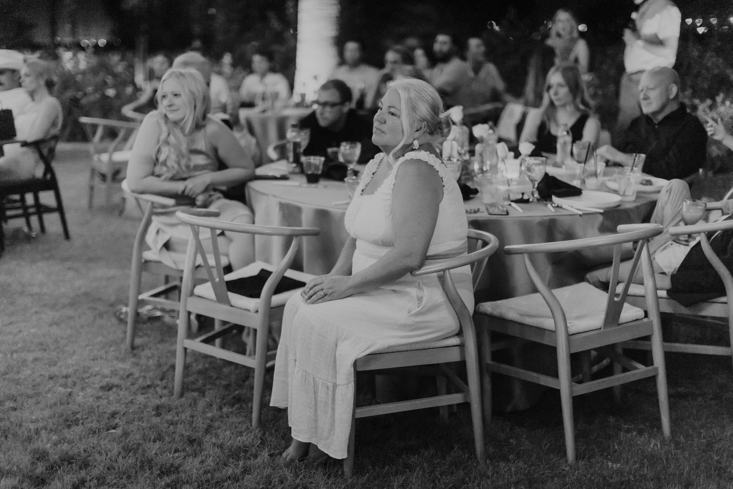 Black and white photograph of an outdoor reception or party gathering with people seated at round tables on a lawn.