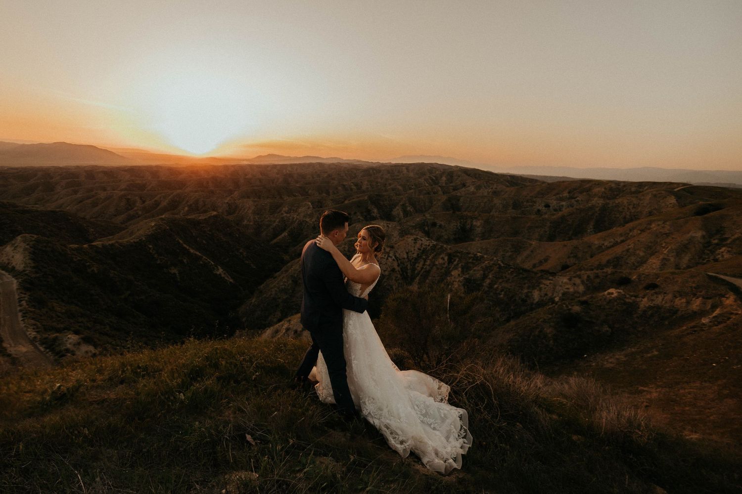 Romantic couple embraces at sunset on mountain cliffs with white wedding dress flowing in the wind.
