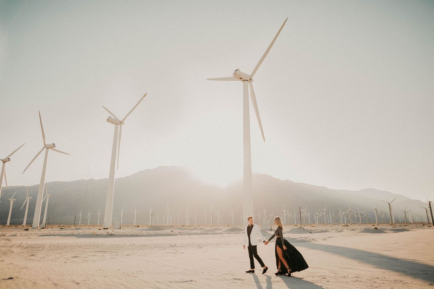 Windmills line the horizon of a desert landscape at sunset with a silhouetted figure in the distance.