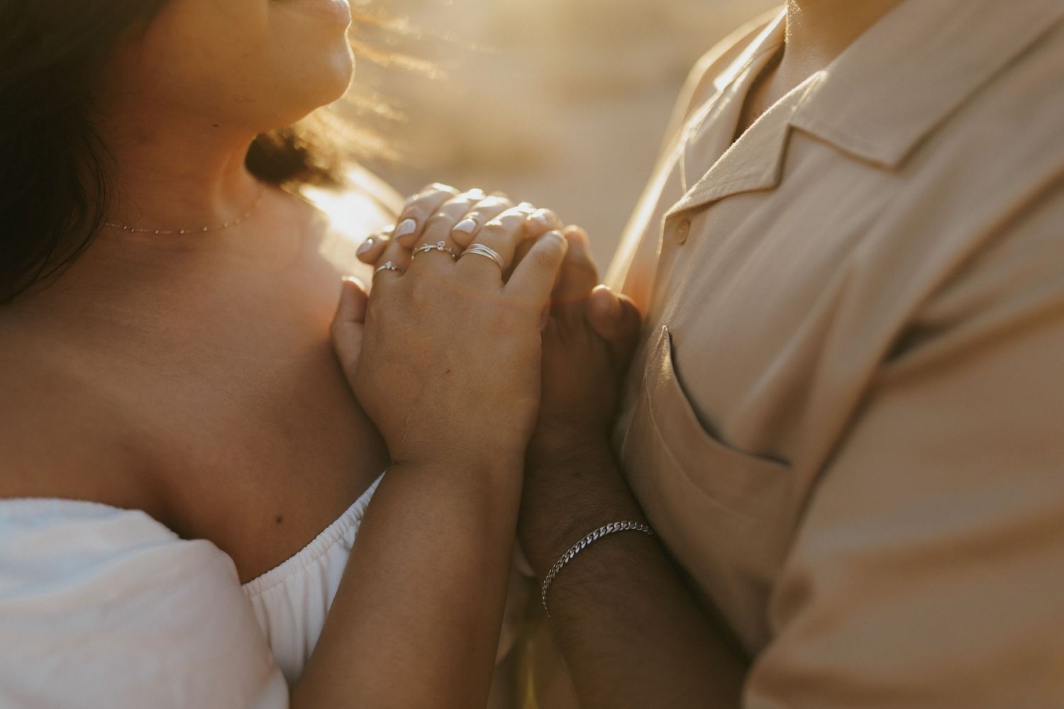 Couple holding hands tenderly in golden sunlight, woman wearing engagement ring and bracelet, romantic outdoor moment.