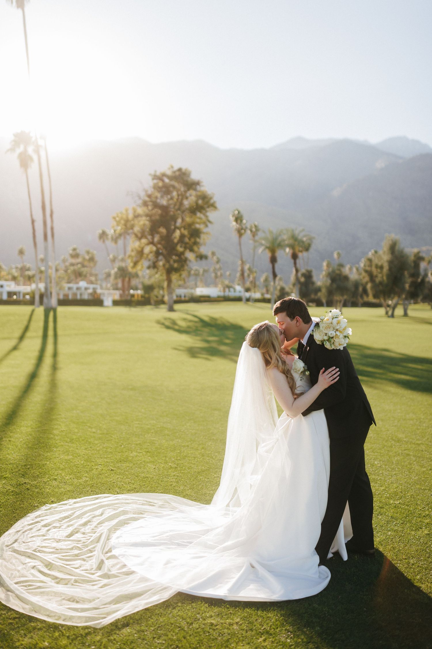 Bride and groom share a kiss on a golf course with palm trees and mountains behind.