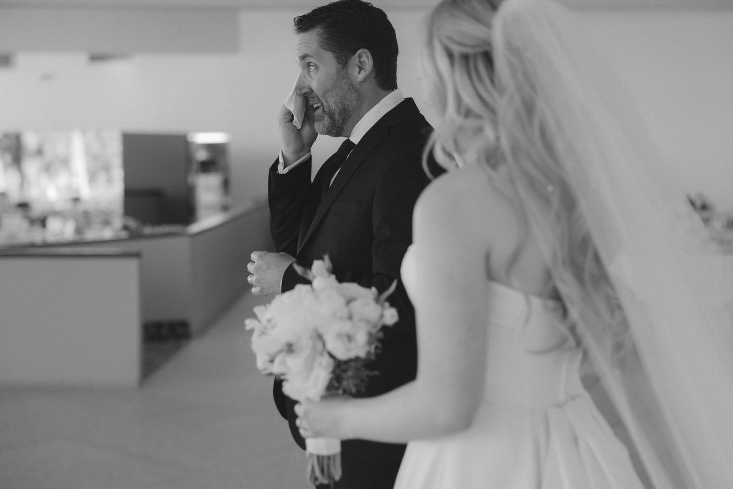 Emotional groom wipes tears as bride holds bouquet in black and white wedding photo.