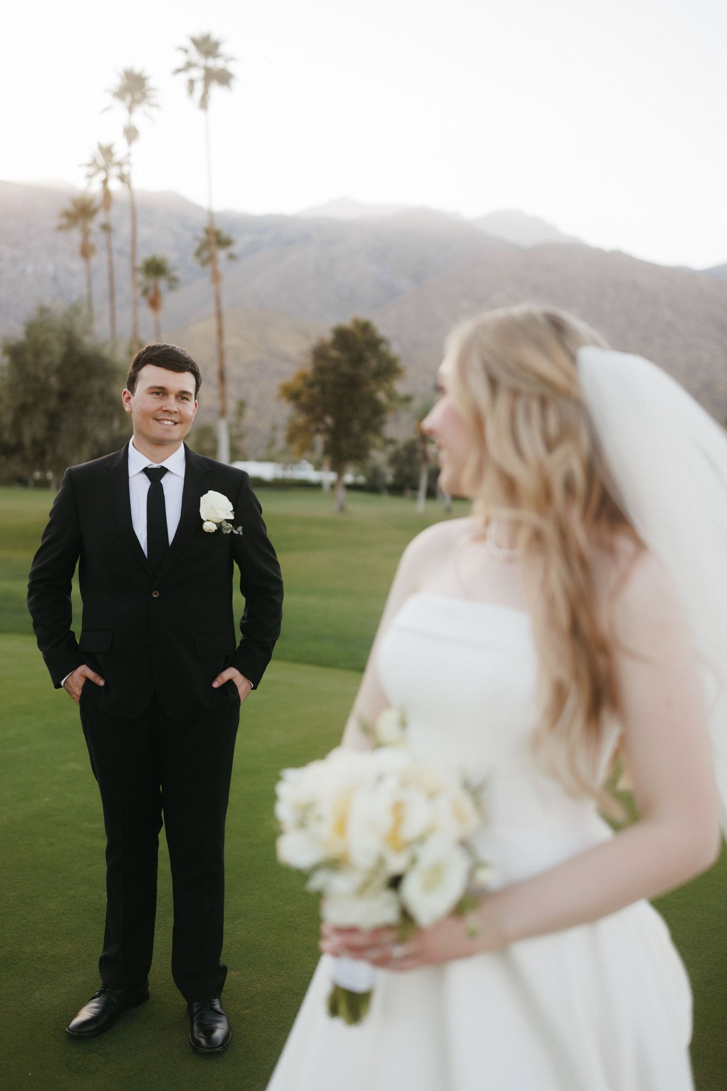 Smiling groom in black suit watches bride holding white bouquet on golf course with mountains.
