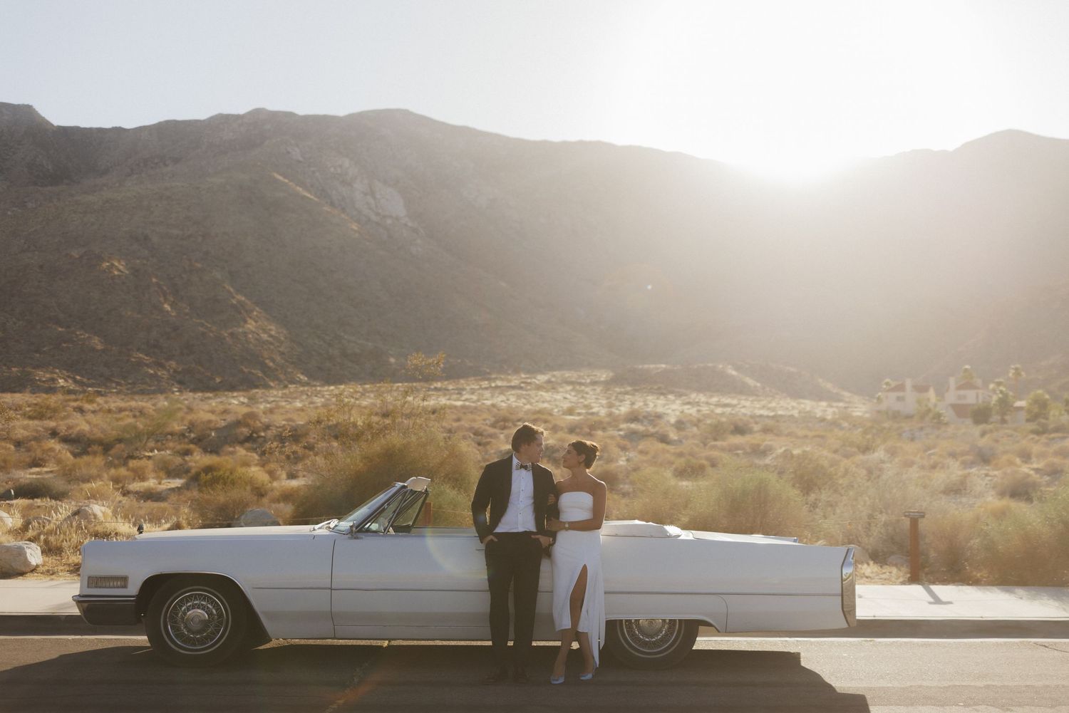 Couple in wedding attire lean against a white vintage convertible with dramatic desert mountains at sunset.