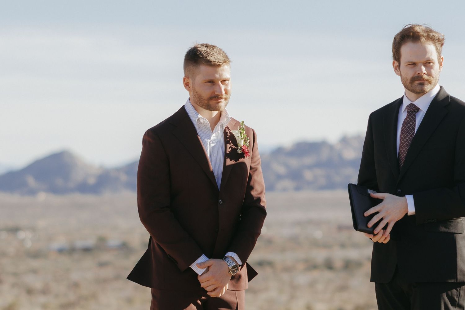 Groom in burgundy suit with boutonniere stands outdoors at desert wedding ceremony with officiant.