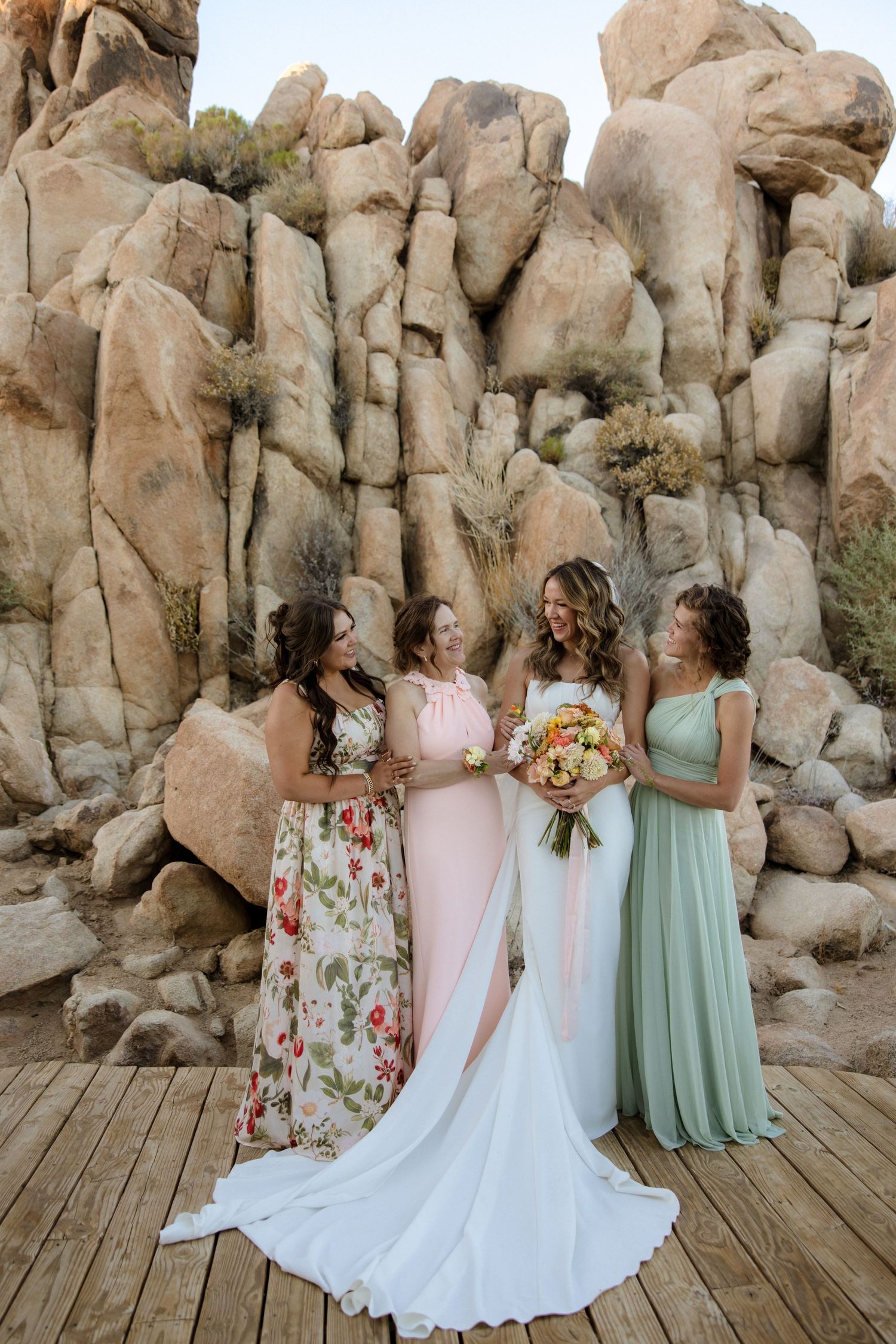 Wedding party wearing floral and pastel dresses pose together against dramatic desert rock formations in natural lighting.