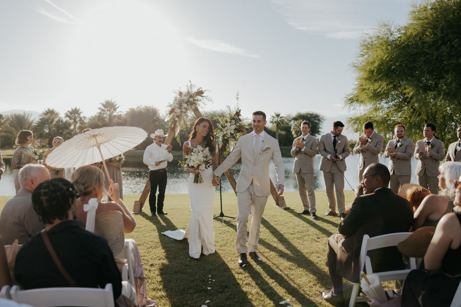 Wedding party celebrates on a sunlit lawn with parasol and guests during an outdoor ceremony.