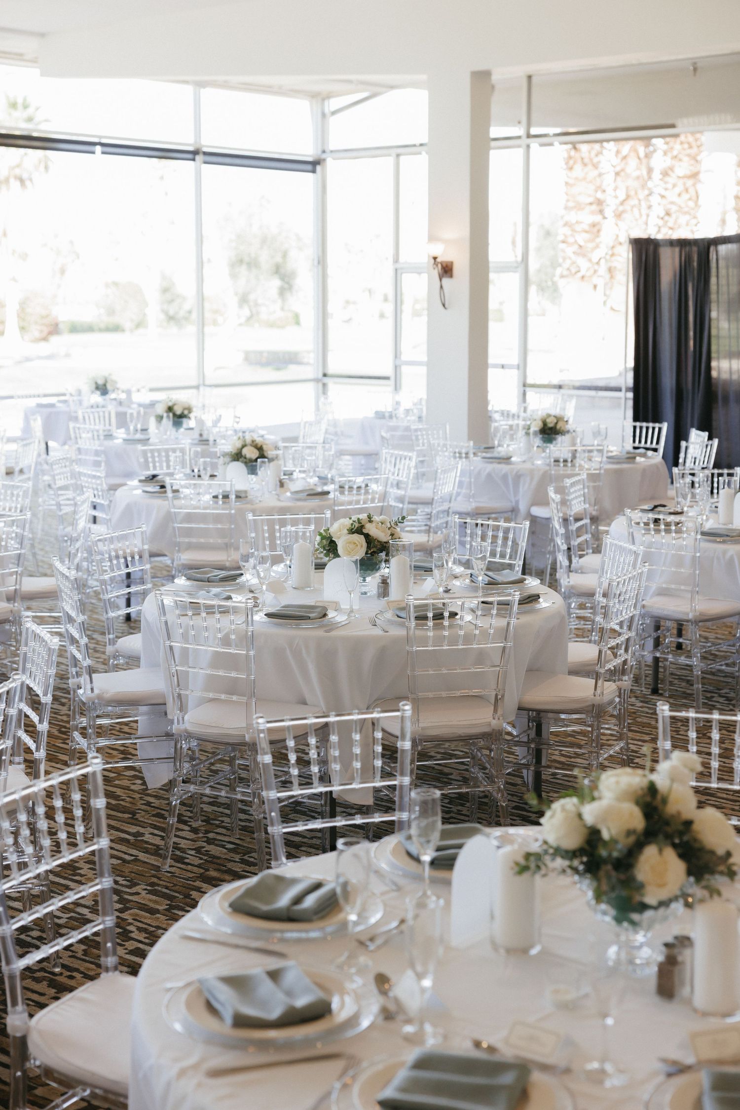 Elegant wedding reception with white round tables, clear chiavari chairs, and white floral centerpieces in a bright venue.