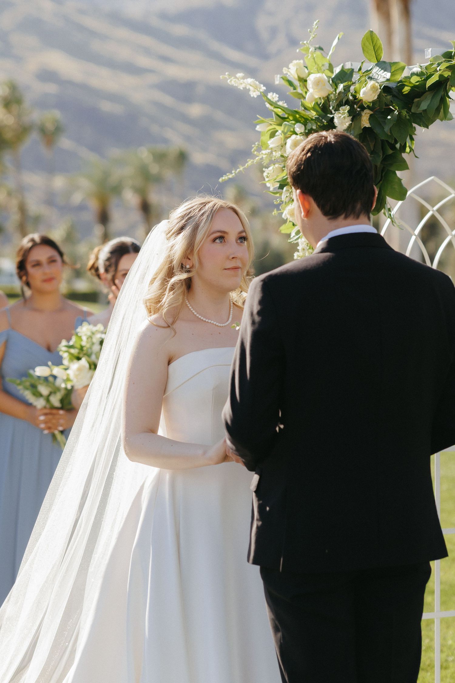 Bride in white gown and veil exchanges vows with groom at outdoor wedding ceremony.