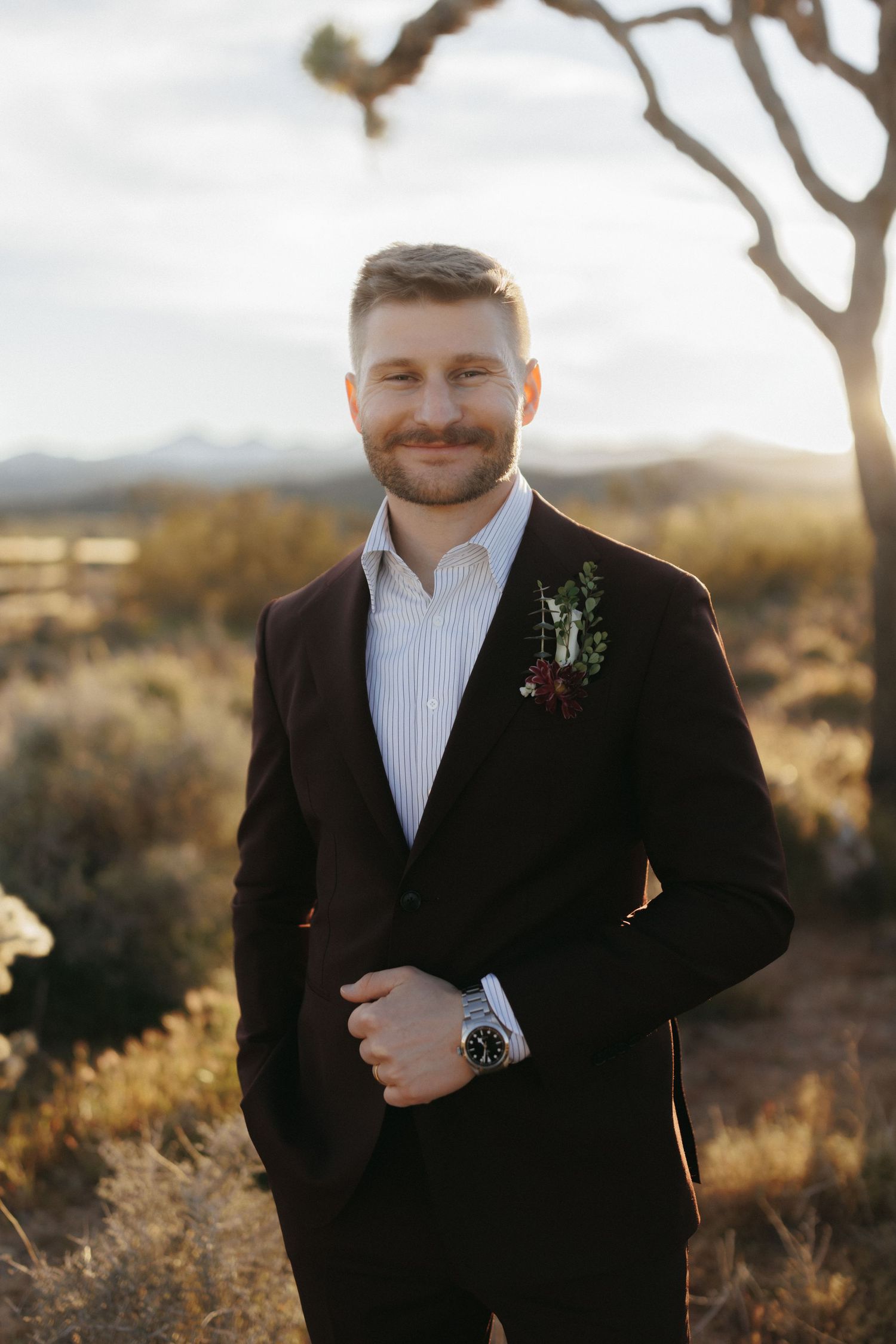 Smiling groom in dark suit with floral boutonniere standing outdoors in desert landscape at golden hour.