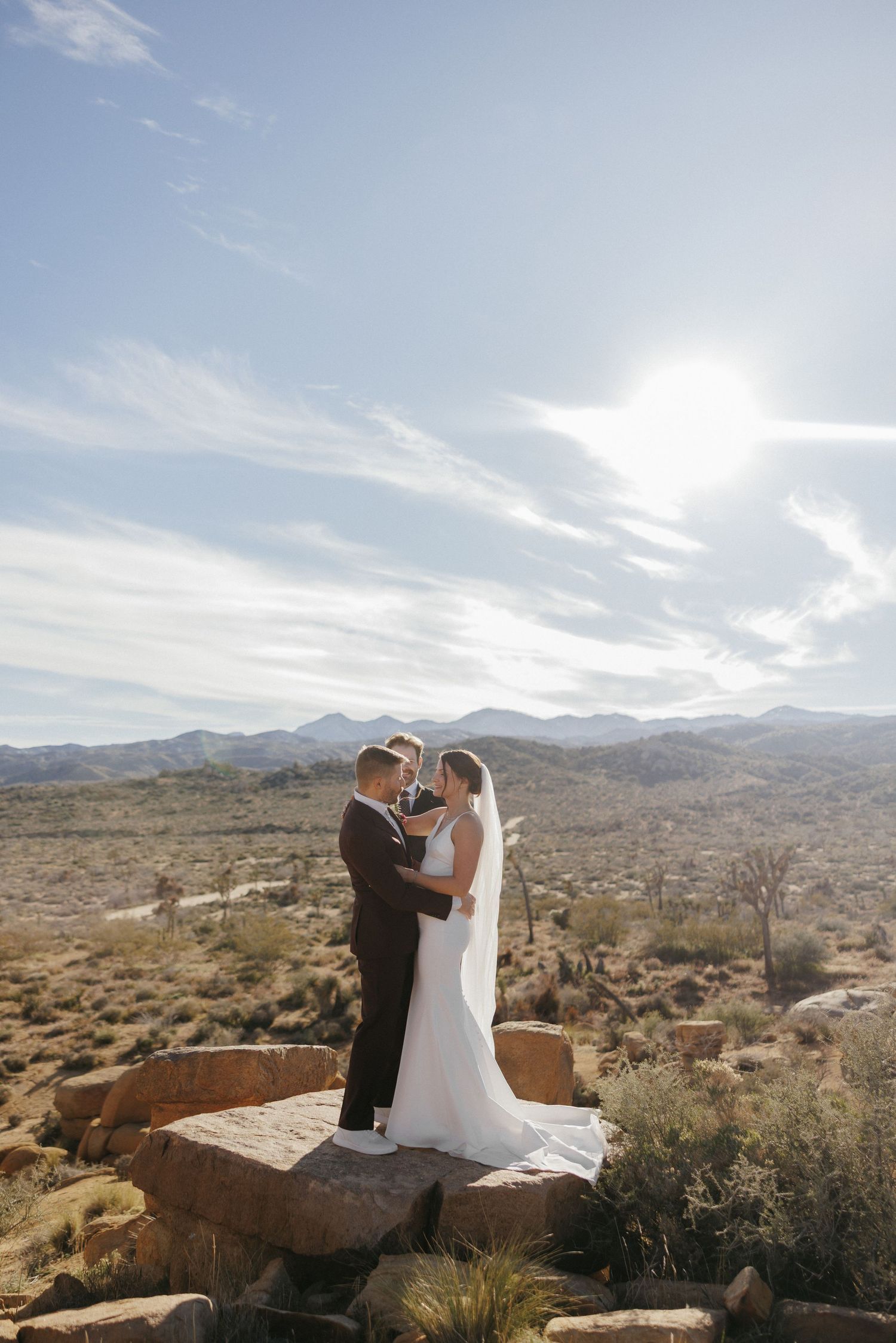 Bride and groom stand on desert rocks at Joshua Tree, sharing an intimate moment with vast landscape and blue sky behind them.