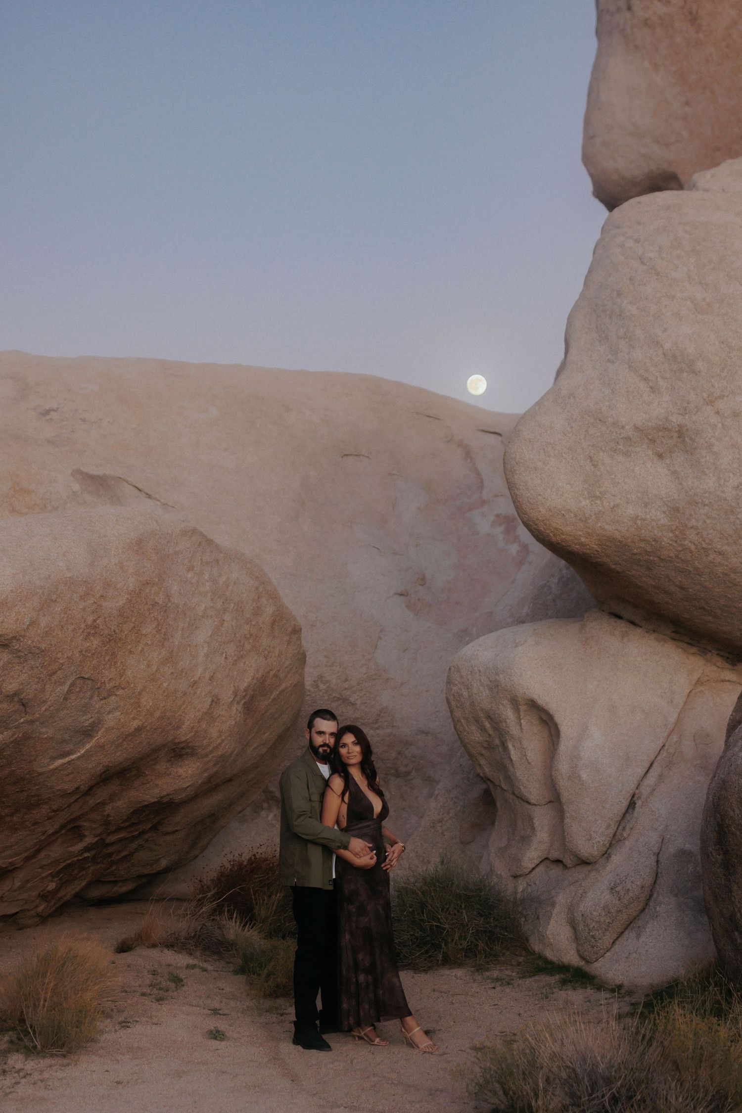 Romantic couple embraces among giant boulders under a full moon at dusk in Joshua Tree National Park.
