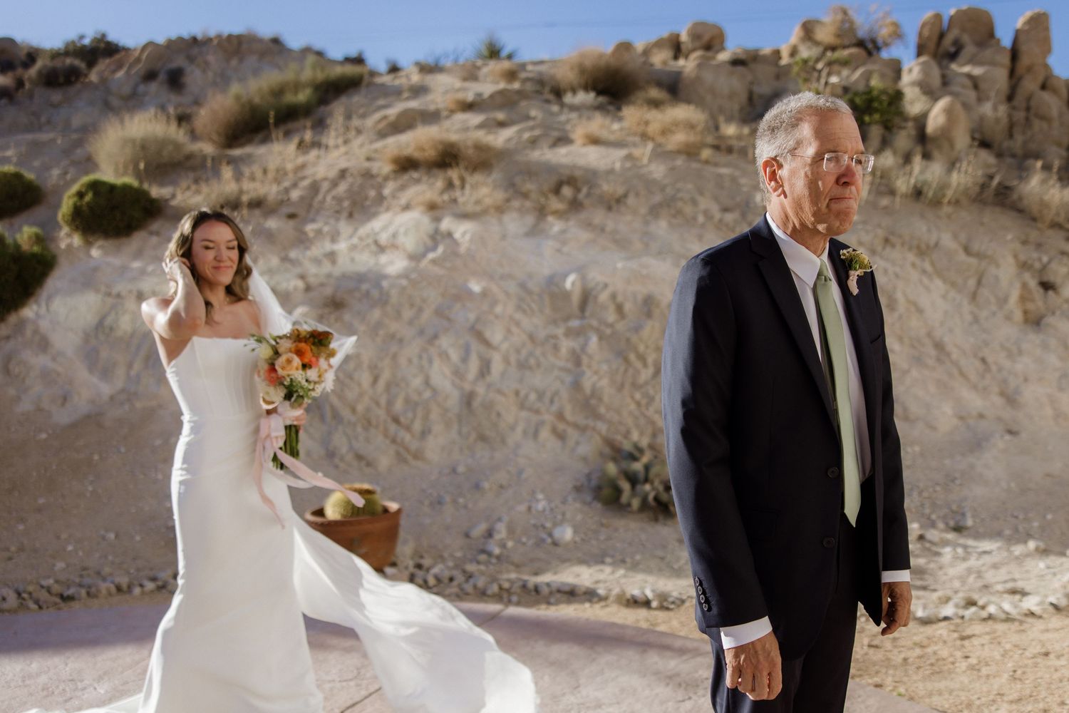 Wedding couple walks hand in hand along rocky desert landscape in formal attire with beautiful mountain backdrop.