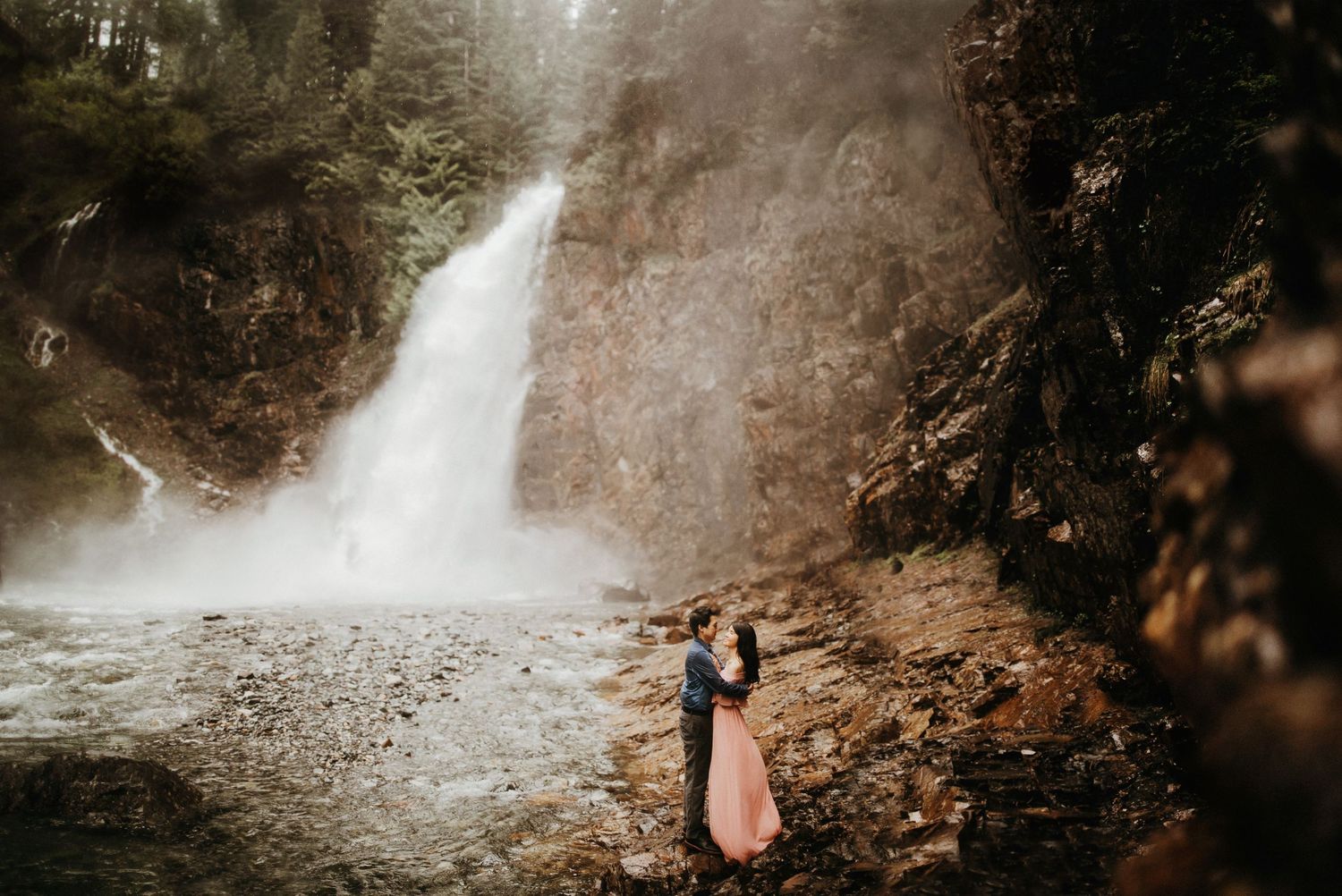 A couple embraces near a misty waterfall cascading down rocky cliffs.