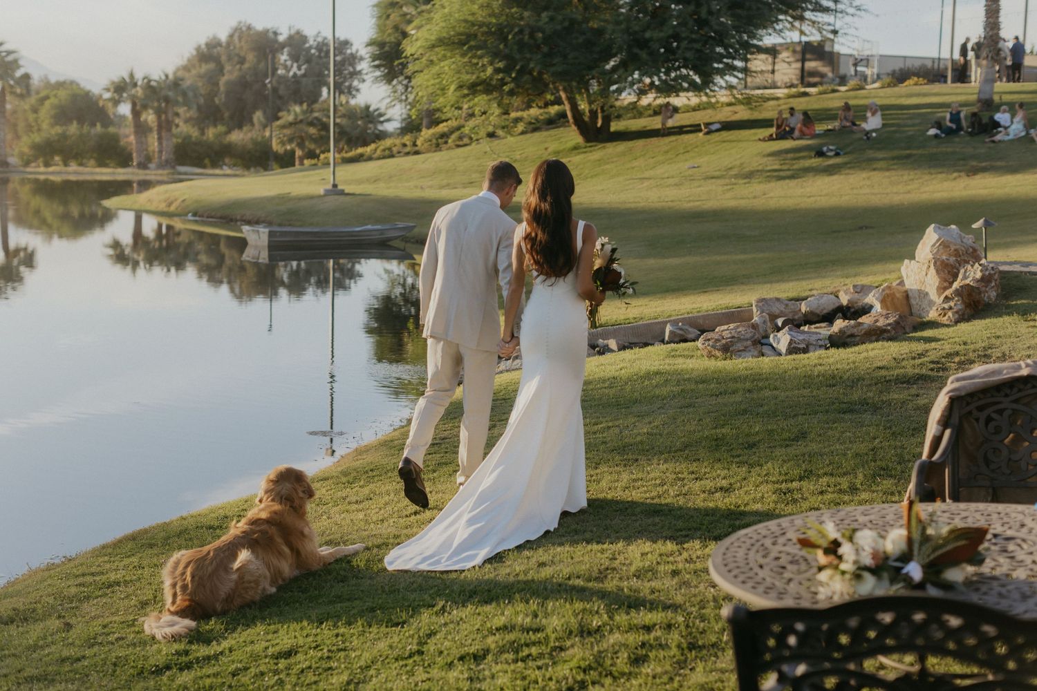 A couple in wedding attire stands by a serene lake as a dog rests on the grass during golden hour.