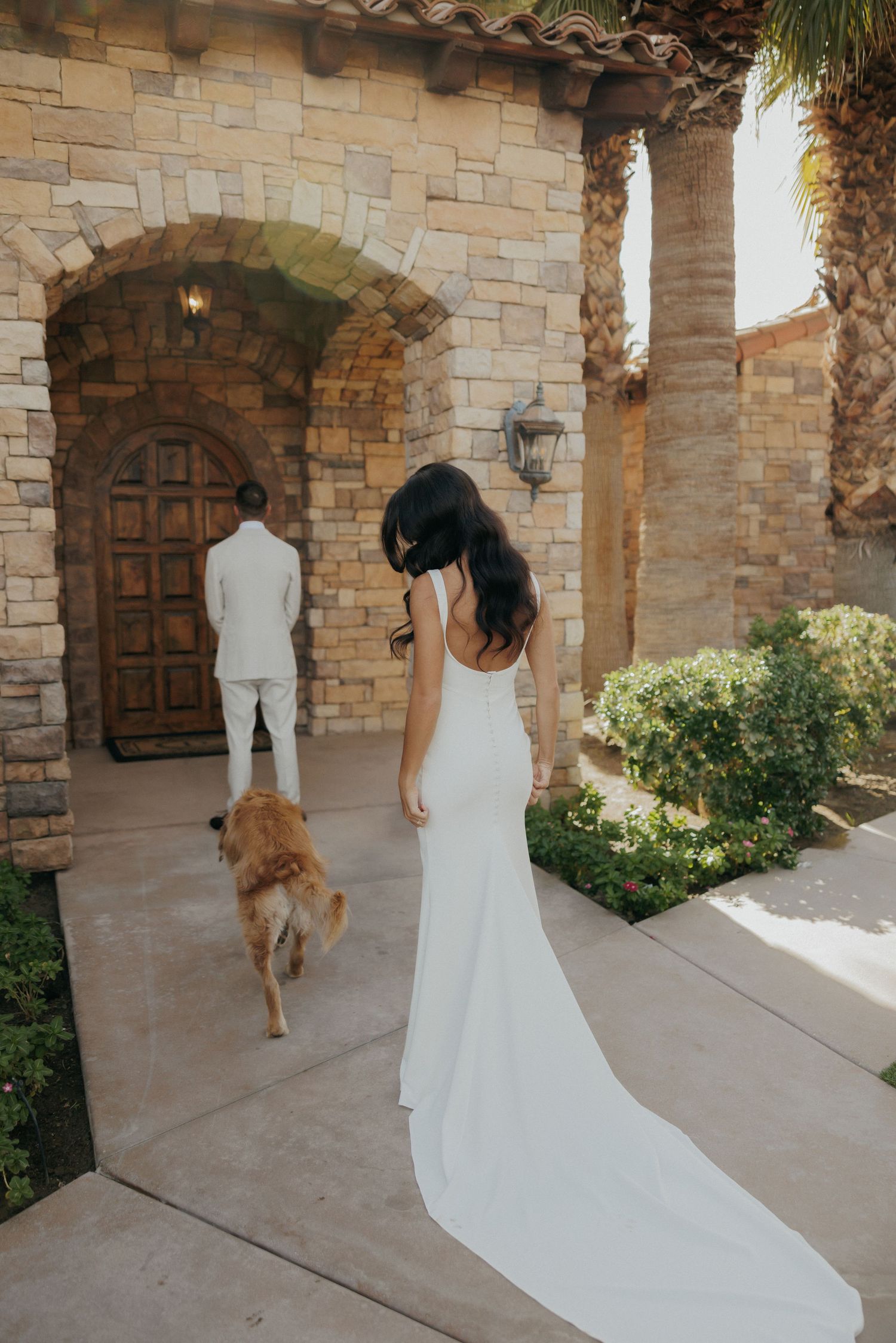 A white wedding dress with a flowing train is photographed hanging in a beautiful stone archway with palm trees.