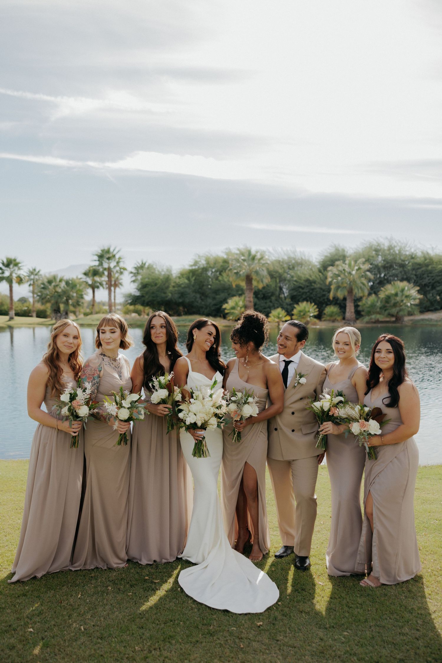 A wedding party in taupe bridesmaid dresses poses together on a grassy lawn beside a waterfront view at sunset.