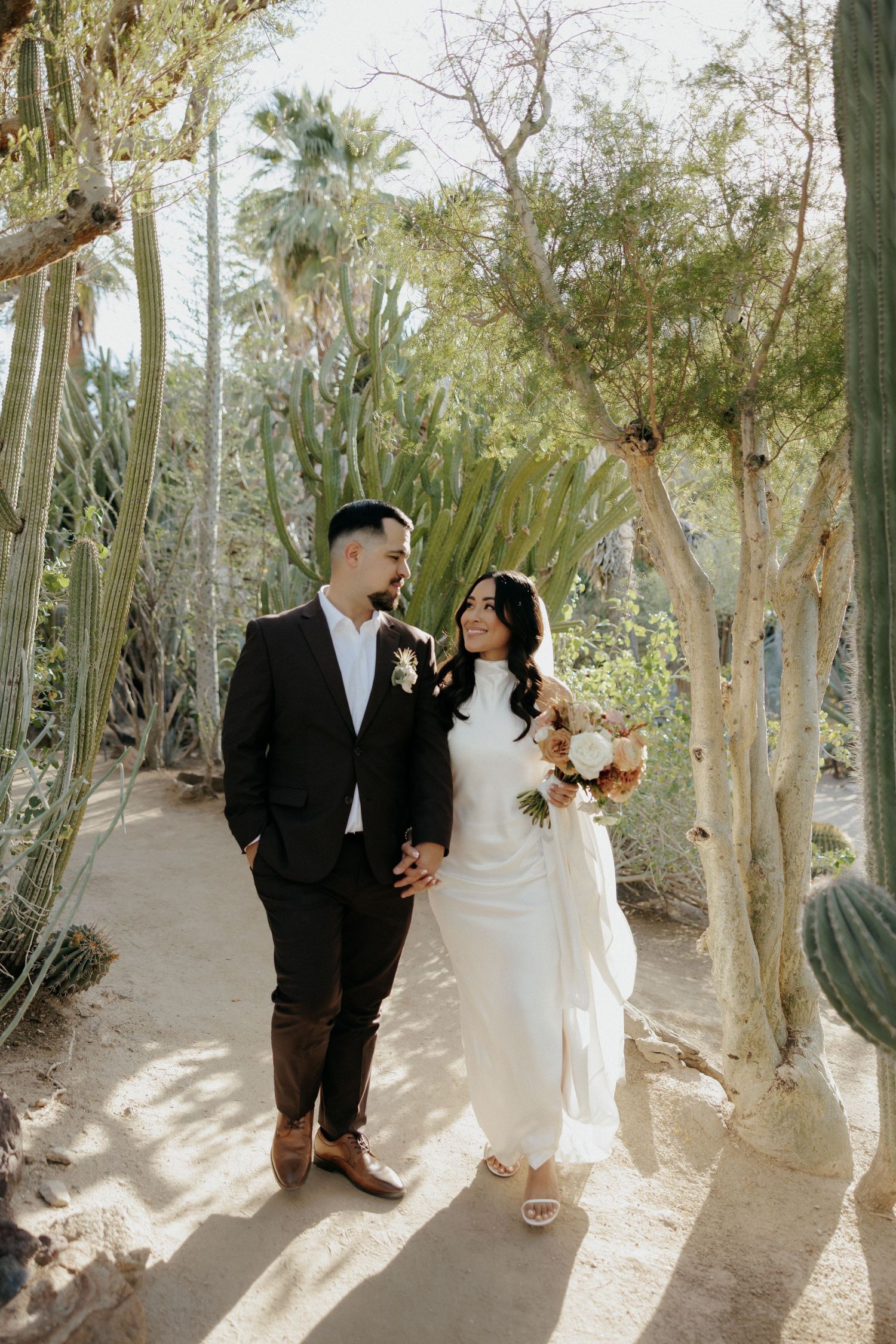 Newlywed couple poses in a desert garden setting surrounded by tall cacti and palm trees in natural sunlight.