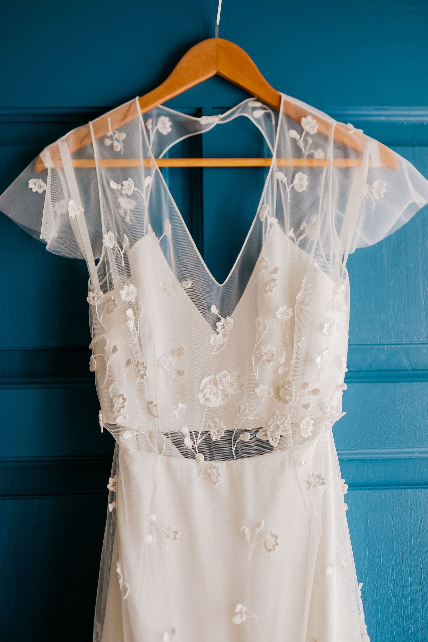 A delicate white wedding dress with floral lace details hangs on a wooden hanger against a blue wall.