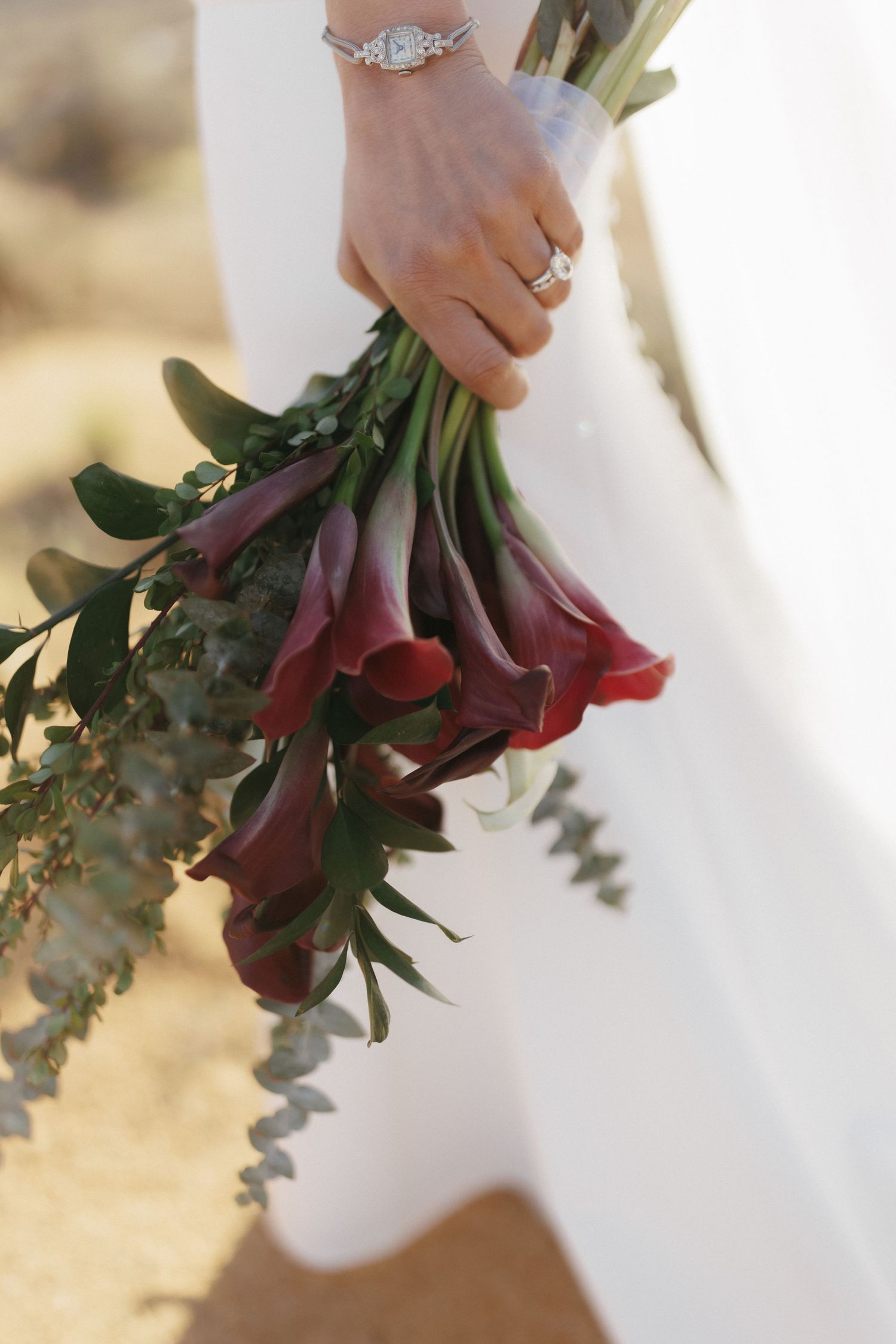 Bride holds elegant bouquet of deep red calla lilies and eucalyptus with diamond ring and bracelet.