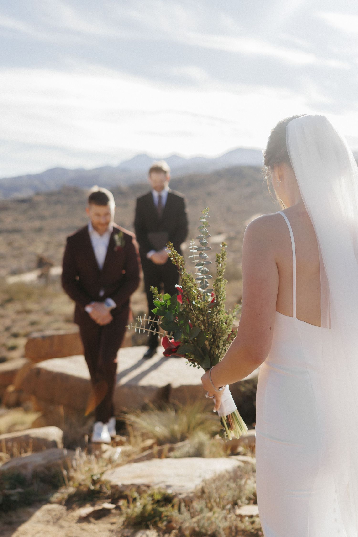 Bride in white gown holding wildflower bouquet walks toward groom in dark suit at outdoor desert wedding ceremony.
