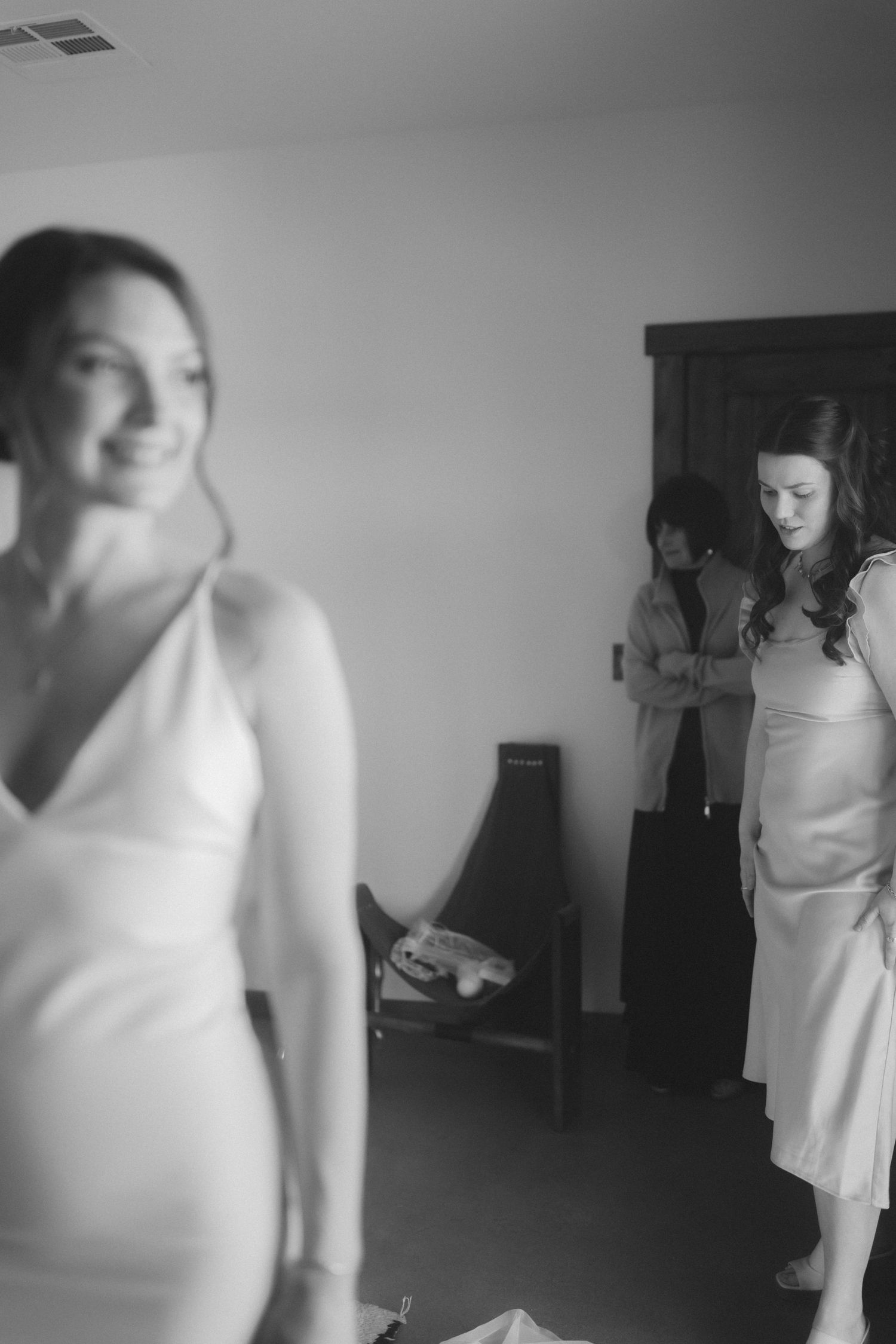 Black and white photo of bride in white gown smiling while bridesmaids prepare in background.
